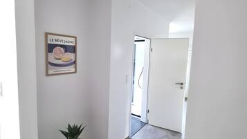 Ein Flur mit einer Tür zu einem hellen Raum, einer Topfpflanze und einem gerahmten Schild an der Wand.