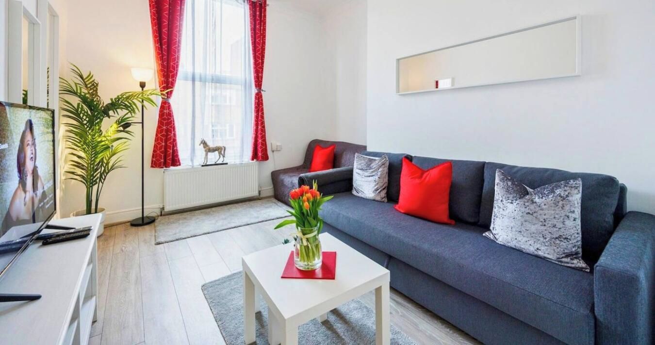 A modern living room with a grey sofa, red and grey cushions, a small white coffee table with a vase of flowers, a television, and a plant.