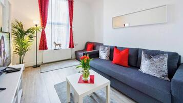 A modern living room with a grey sofa, red and grey cushions, a small white coffee table with a vase of flowers, a television, and a plant.