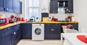 A compact kitchen with a washing machine, a coffee maker, and a toaster. - 1BR | Sleeps 5 | 1 min to Tube | West Kensington (Greater London)