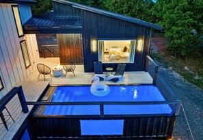 A hot tub with a swan float, a deck with chairs, and a view of a bedroom with a bed and curtains. - King Suite | Mtn Views | EV Charger | Heated Pool (Broken Bow)