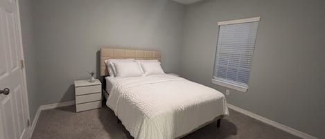 A bedroom with a bed, a nightstand, a ceiling fan, a window with blinds, and a door.