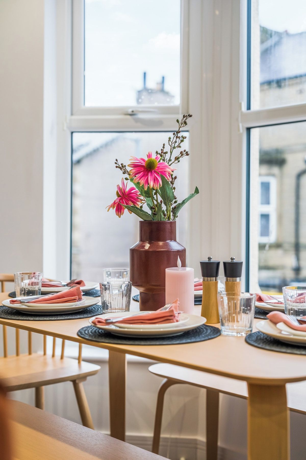 A dining table set with plates, glasses, and a centerpiece of pink flowers in a brown vase.