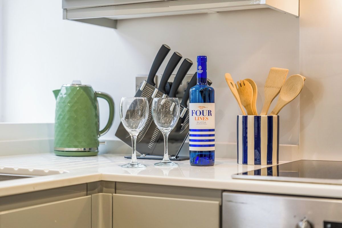 A kitchen counter with a green kettle, four wine glasses, a bottle of Four Lines Rueda wine, and wooden utensils.