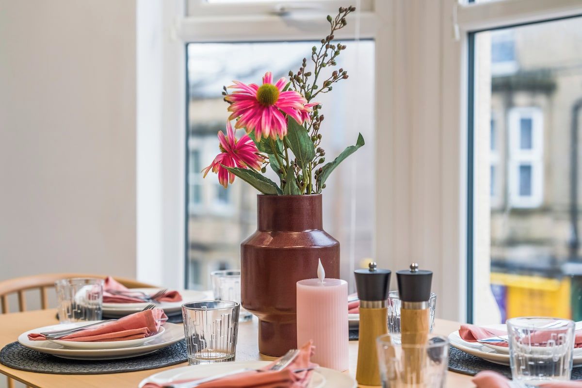 Una mesa de comedor dispuesta con platos, cubiertos, vasos, un florero con flores, una vela y saleros y pimenteros.
