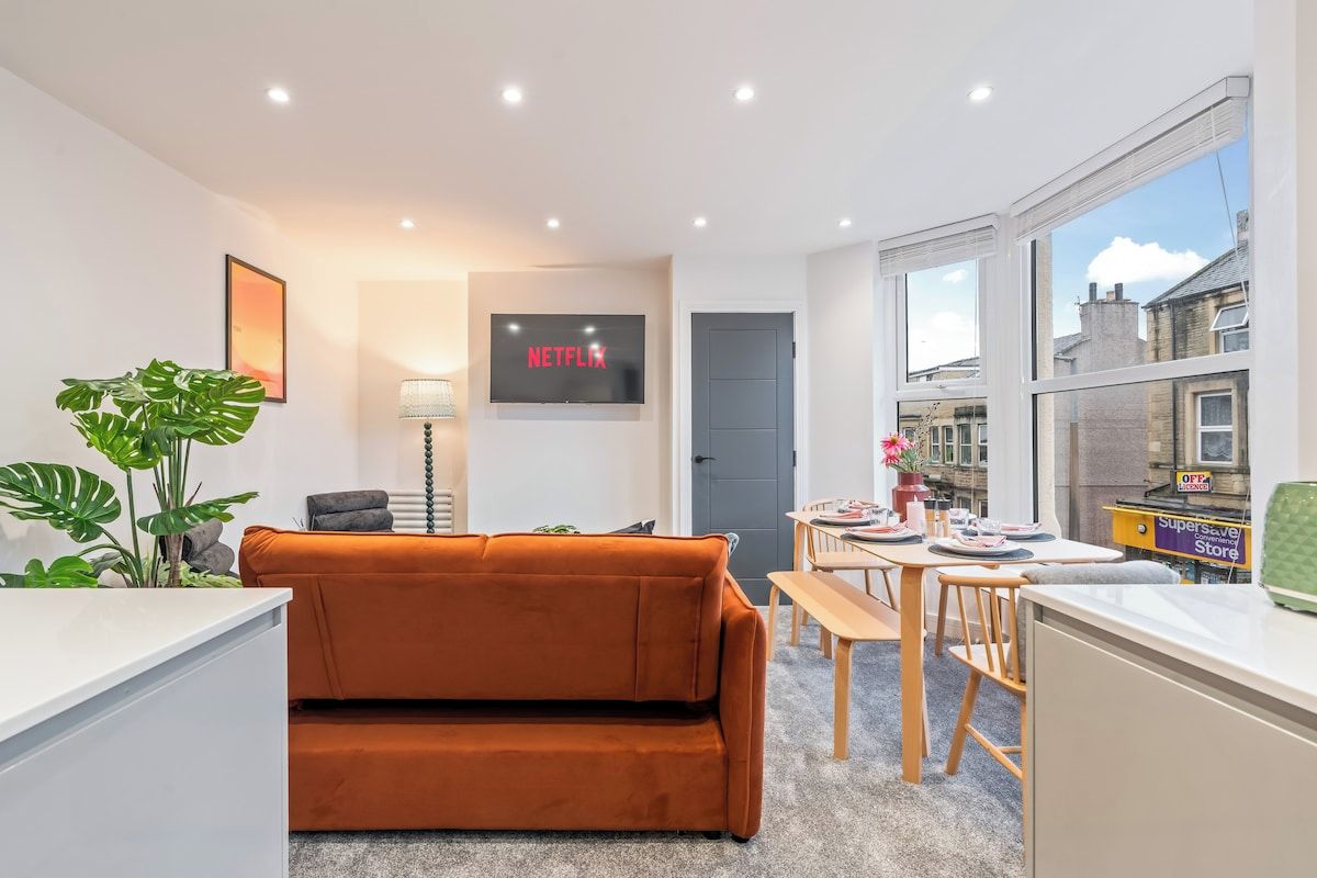 A modern living room with a sofa, a television displaying 'NETFLIX,' a dining area with a table and chairs, and a view of the city through large windows.