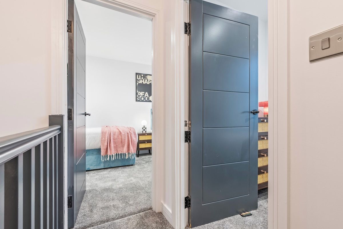 A bedroom with a bed, a nightstand, a wall-mounted shelf, and a door leading to another room.