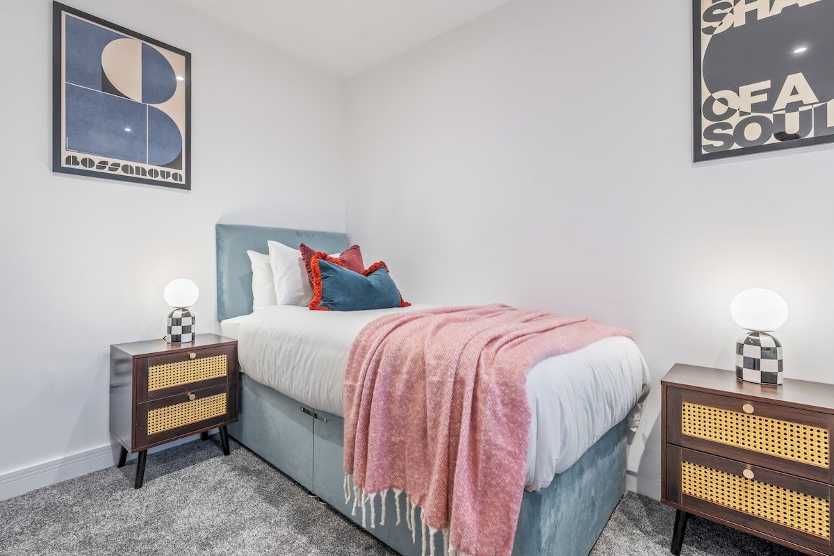 A bedroom with a bed, two bedside tables with lamps, a wall art piece, and a carpeted floor.