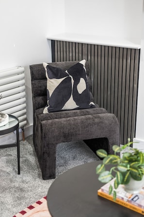 A modern living room with a grey upholstered armchair, a black and white patterned throw pillow, a round coffee table, and a potted plant. - Coastal retreat near Morecambe Bay and promenade (Heysham)