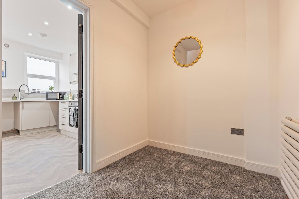 A room with a carpeted floor, a white wall with a gold-framed mirror, and a door leading to a kitchen area.