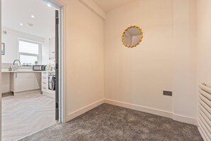 A room with a carpeted floor, a white wall with a gold-framed mirror, and a door leading to a kitchen area. - Coastal retreat near Morecambe Bay and promenade (Heysham)