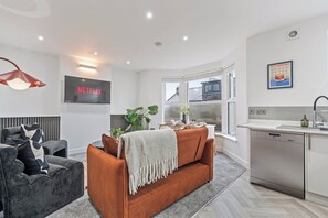 A modern living room with a grey sofa, a brown sofa, a TV displaying 'NETFLIX', and a kitchen area with a dishwasher. - Coastal retreat near Morecambe Bay and promenade (Heysham)