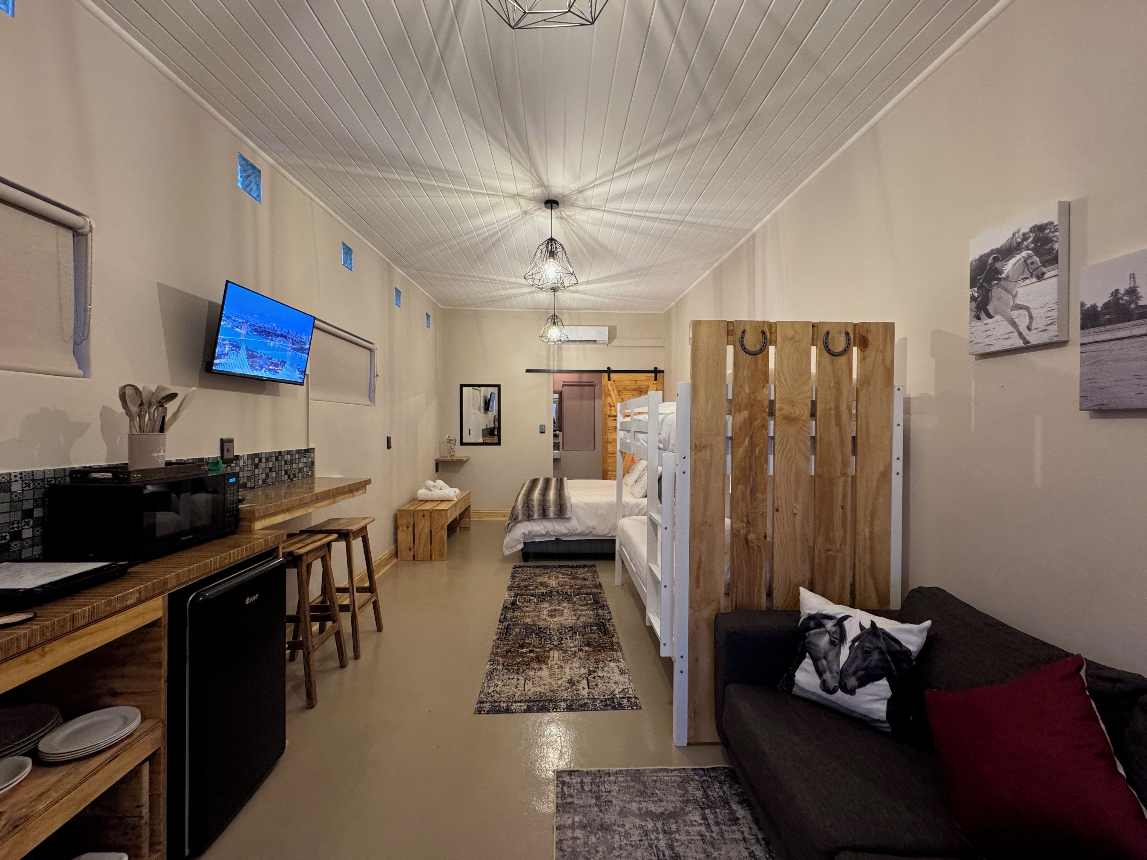 Una habitación de hotel moderna con kitchenette, una cama con cabecera de madera, un televisor y un comedor con microondas y taburetes.