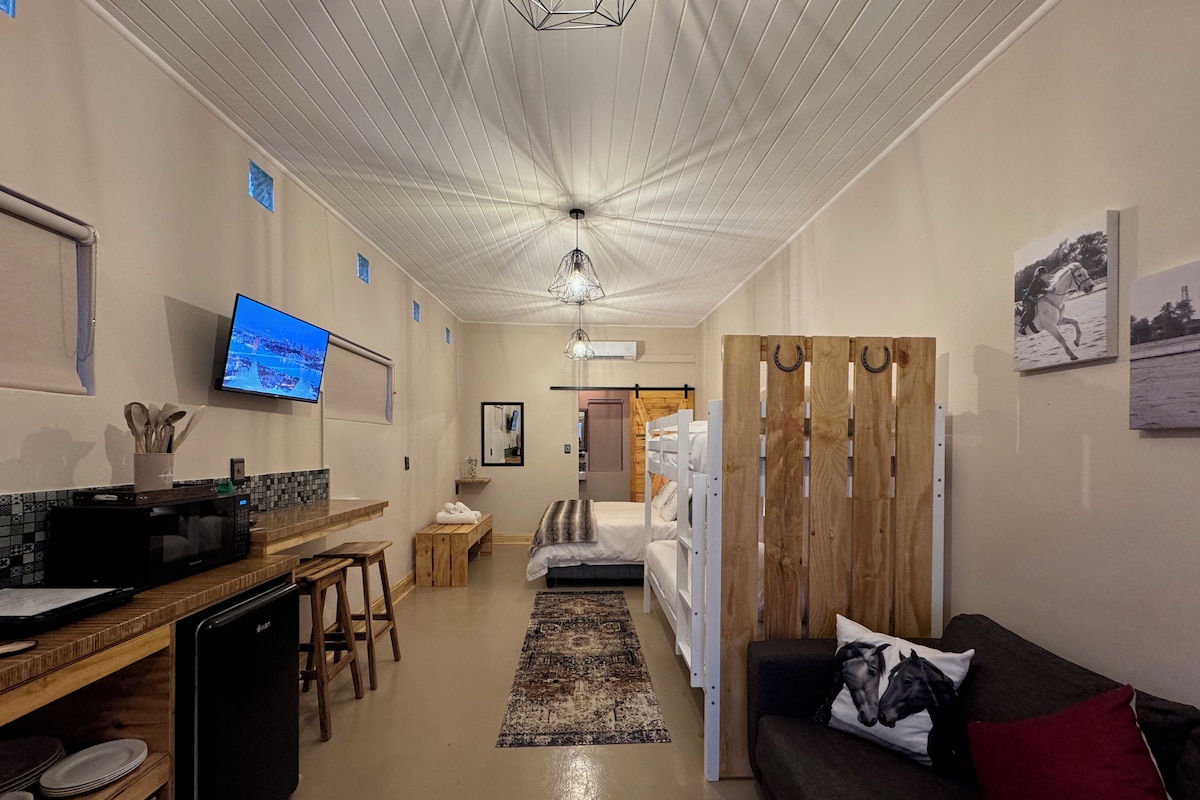 A modern hotel room with a kitchenette, a bed with a wooden headboard, a TV, and a dining area with a microwave and stools.