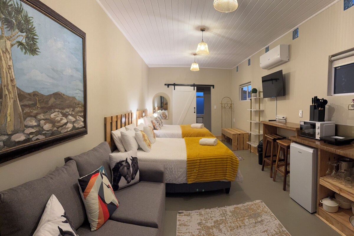 A hotel room with a bed, a sofa, a desk with a computer, a TV, and a painting on the wall.
