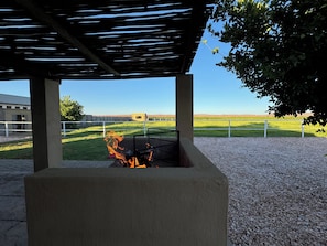 A covered outdoor area with a fire pit, a view of a building in the background, and a fenced field.