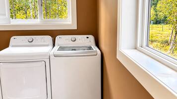 A laundry room with two white washing machines, a window with a view of trees, and brown walls.