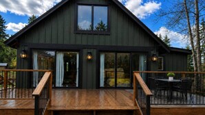 A two-story wooden cabin with a deck, large windows, and a staircase leading to the entrance. - Beaver Creek Chalet | 1 Bedroom | Sleeps 6 | Pet Friendly (Alberni-Clayoquot)