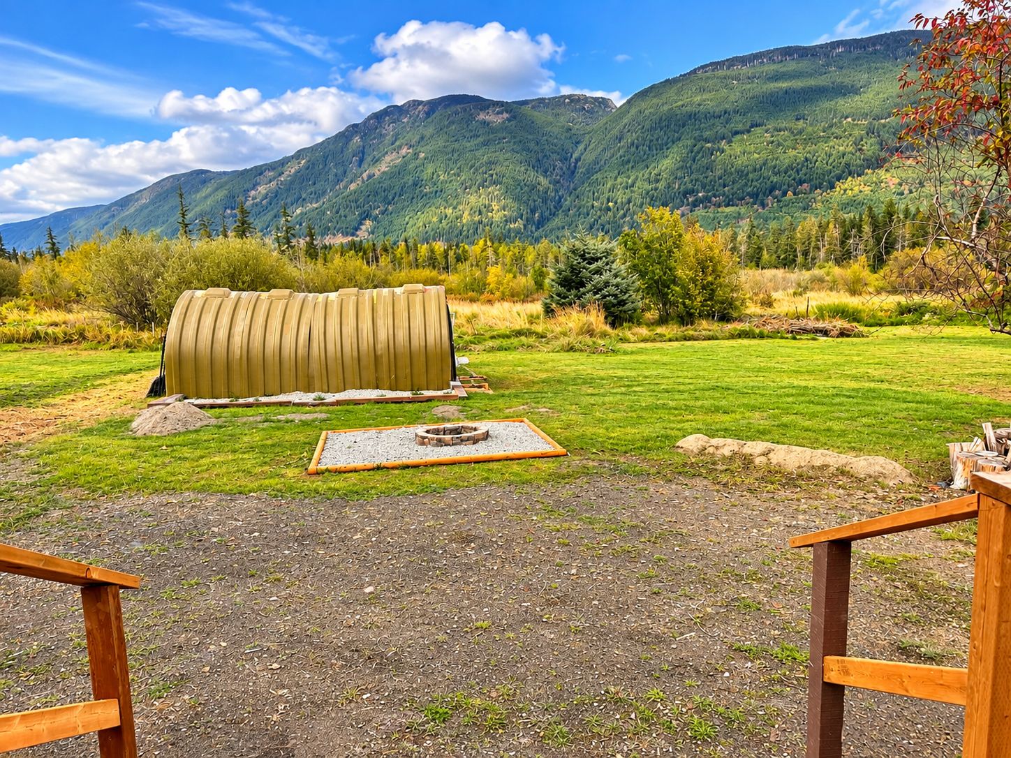 Un espace extérieur rustique aménagé avec un banc en bois, un foyer et un grand objet cylindrique, le tout se détachant sur un arrière-plan de collines verdoyantes et d’un ciel dégagé.