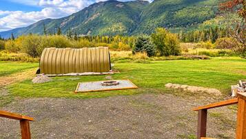 Un espace extérieur rustique aménagé avec un banc en bois, un foyer et un grand objet cylindrique, le tout se détachant sur un arrière-plan de collines verdoyantes et d’un ciel dégagé.