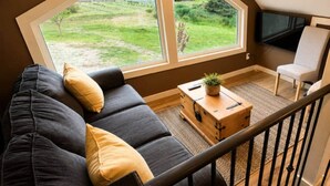 A modern living room with a dark sofa, a wooden coffee table, and a large window offering a view of a grassy area and a small building. - Beaver Creek Chalet | 1 Bedroom | Sleeps 6 | Pet Friendly (Alberni-Clayoquot)