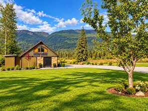 A two-story wooden cabin with a stone facade, surrounded by a well-maintained lawn and a large tree.