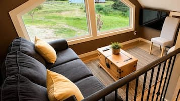 Una sala moderna con un sofá oscuro, una mesa de centro de madera y un amplio ventanal con vistas a una zona verde y un edificio pequeño.