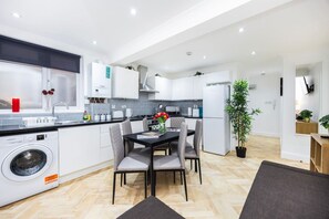 A modern kitchen with white cabinets, a built-in washing machine, and a dining area with a table and chairs. - West Kensington 2BR | Steps to Tube | Sleeps 7 (Greater London)