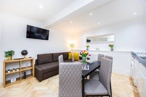 A modern living room with a sofa, a dining table with chairs, a TV mounted on the wall, and a kitchen area with a window. - West Kensington 2BR | Steps to Tube | Sleeps 7 (Greater London)