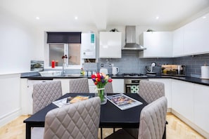 A modern kitchen with white cabinetry, a black countertop, and a dining area with a table and chairs. - West Kensington 2BR | Steps to Tube | Sleeps 7 (Greater London)