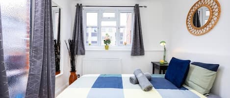 A bedroom with a bed, a window with curtains, a bedside table with a lamp, and a wall-mounted mirror.