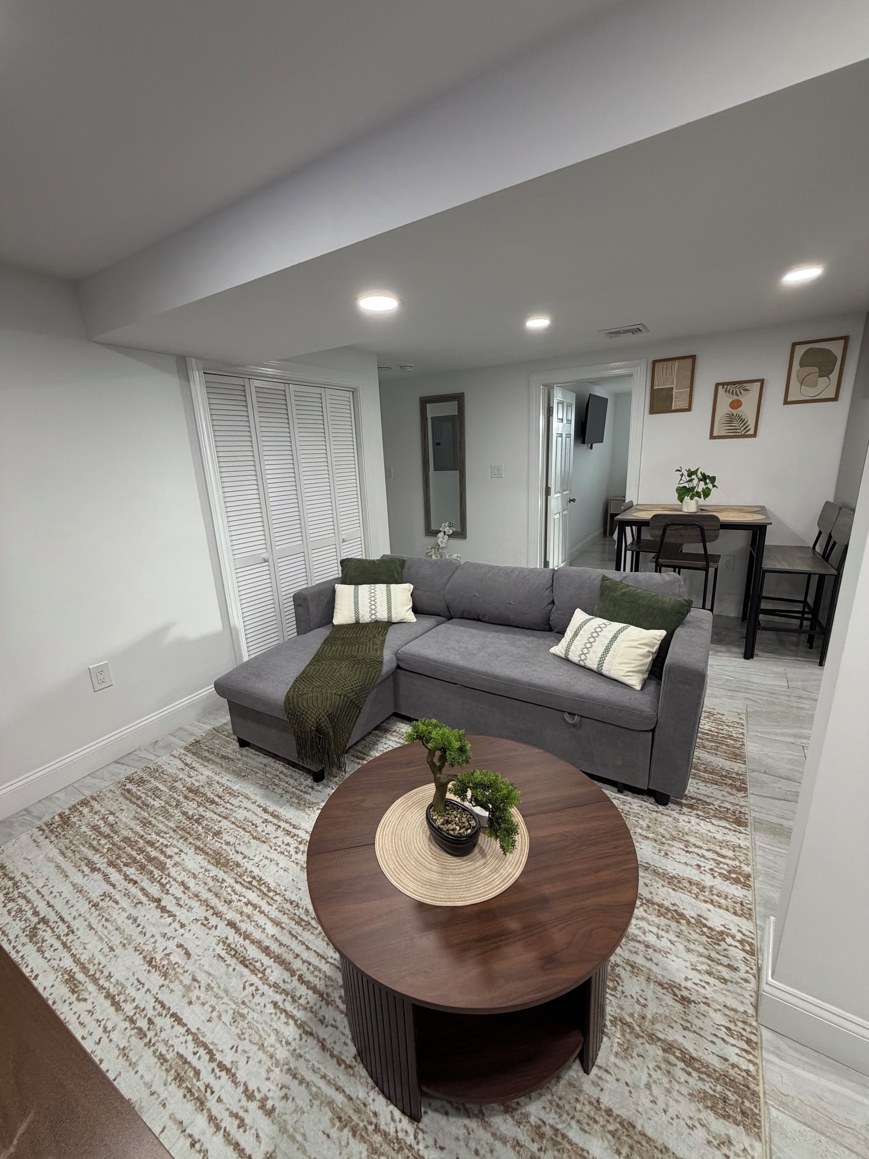 A modern living room with a grey sectional sofa, a round coffee table with a bonsai, and a dining area in the background.