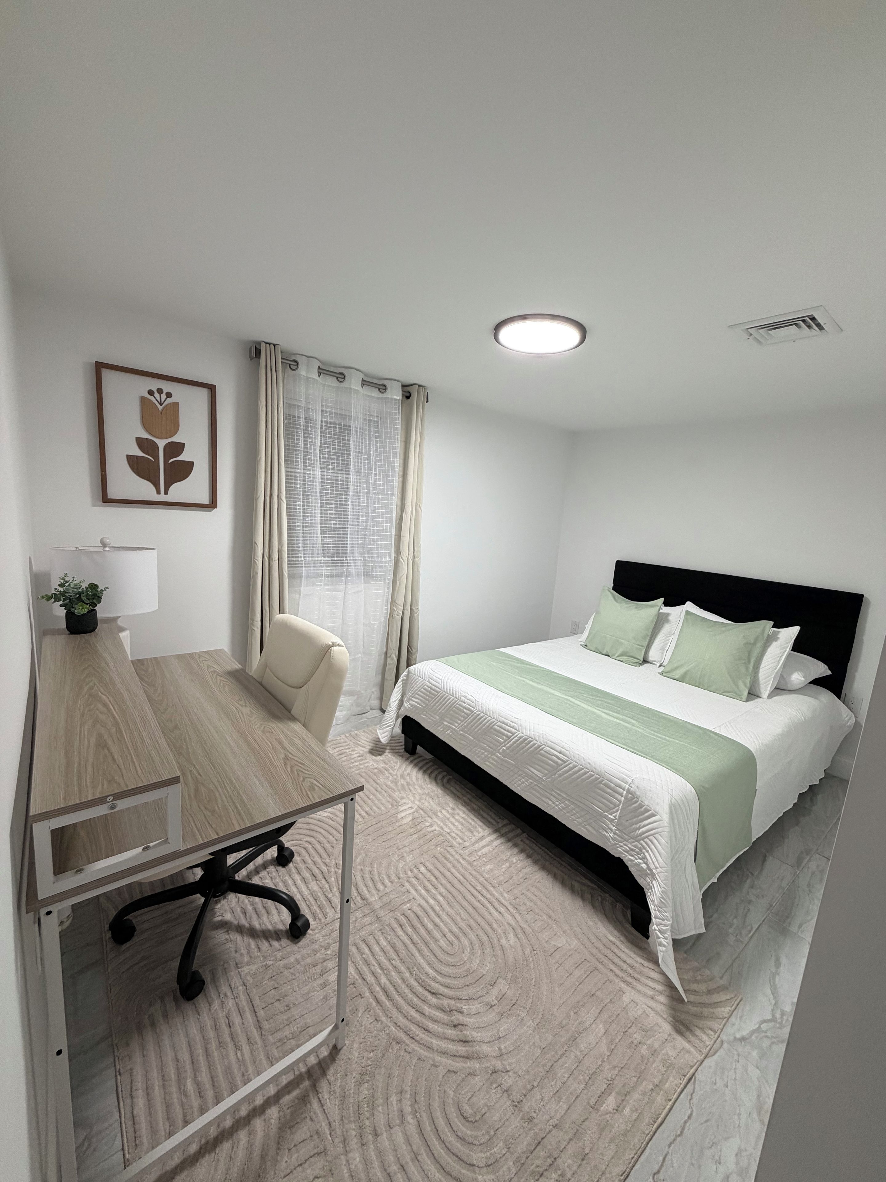 Une chambre d’hôtel avec un lit, un bureau, une chaise, une fenêtre avec des rideaux et un luminaire au plafond.