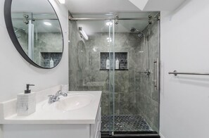 Un baño moderno con ducha envuelta en vidrio, un lavabo blanco con dispensador de jabón y un espejo con marco negro.