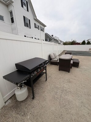 Un patio trasero con una parrilla negra, muebles de mimbre y una valla blanca.