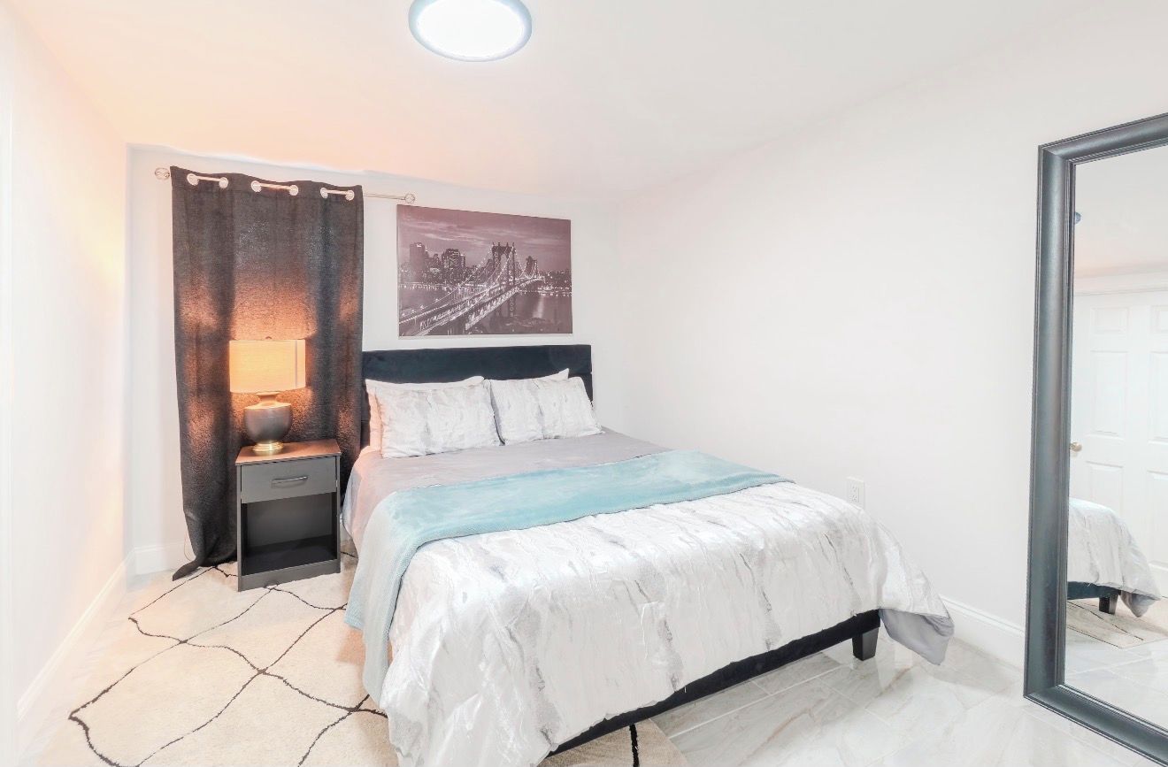 Een slaapkamer met een bed, nachtkastje, lamp en een stadsgezichtschilderij aan de muur.