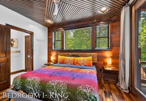 Ein Schlafzimmer mit Holzbalkendecke, einem großen Bett mit bunter Bettdecke, zwei Nachttischlampen und einem Fenster mit Jalousien.