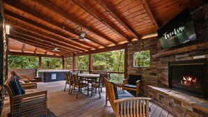 Un espace extérieur abrité doté d’un plancher en bois, d’une cheminée en pierre et d’un écran de télévision diffusant des images de « Tree Line Vacation Rentals ».