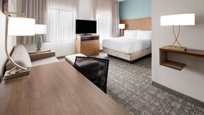 A hotel room with a wooden desk, a television, a bed, a chair, and a nightstand with a lamp. - Accessible King Studio w/ Roll Shower & Breakfast (Harrisburg)