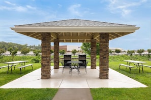 A gazebo with a stone base and a tiled roof, surrounded by picnic tables and a grill area.