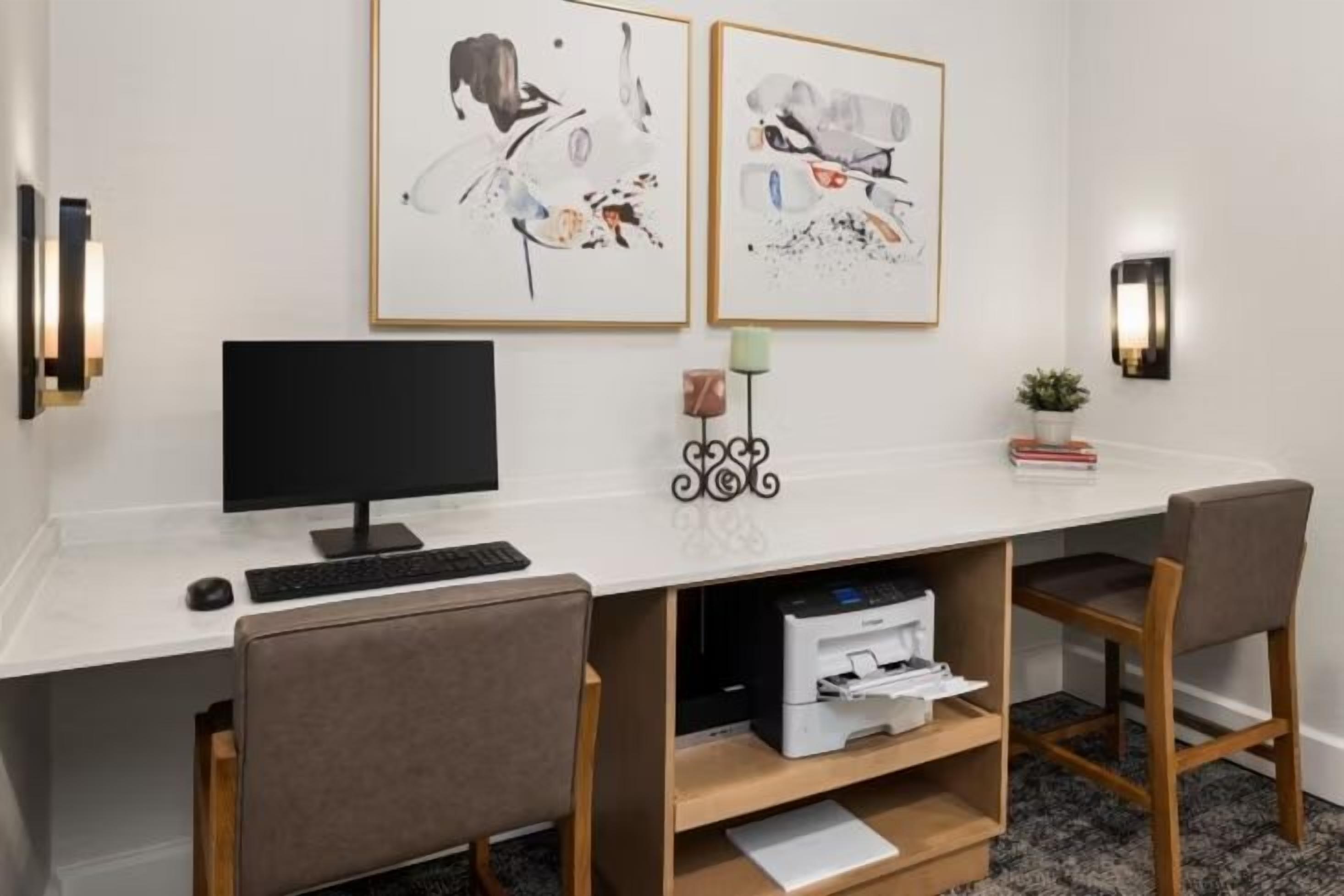 A modern office space with a desk, two chairs, a computer, and framed artwork on the wall.