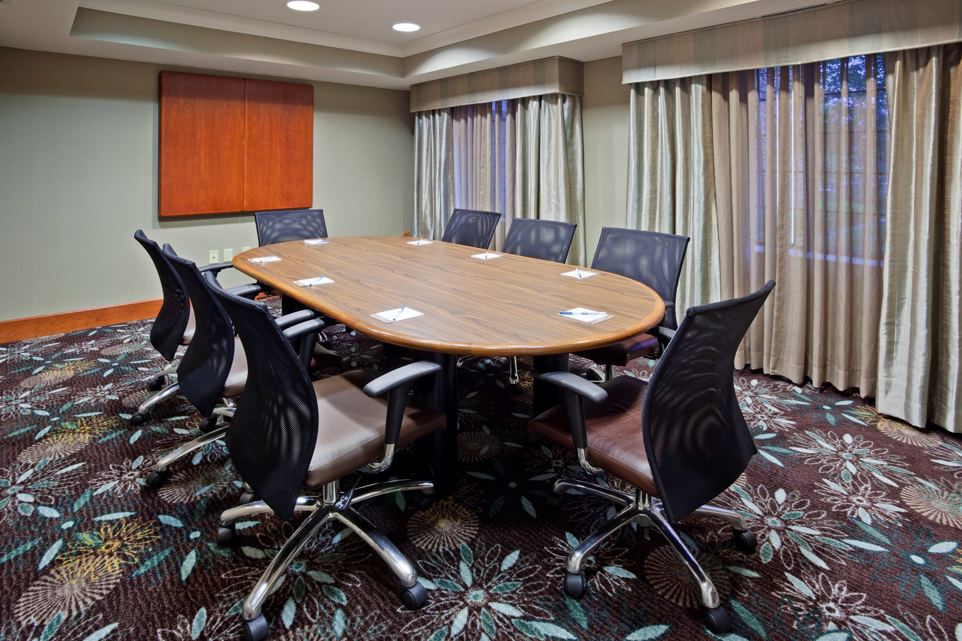 A conference room with a large wooden table surrounded by chairs, a carpeted floor, and curtains on the windows.