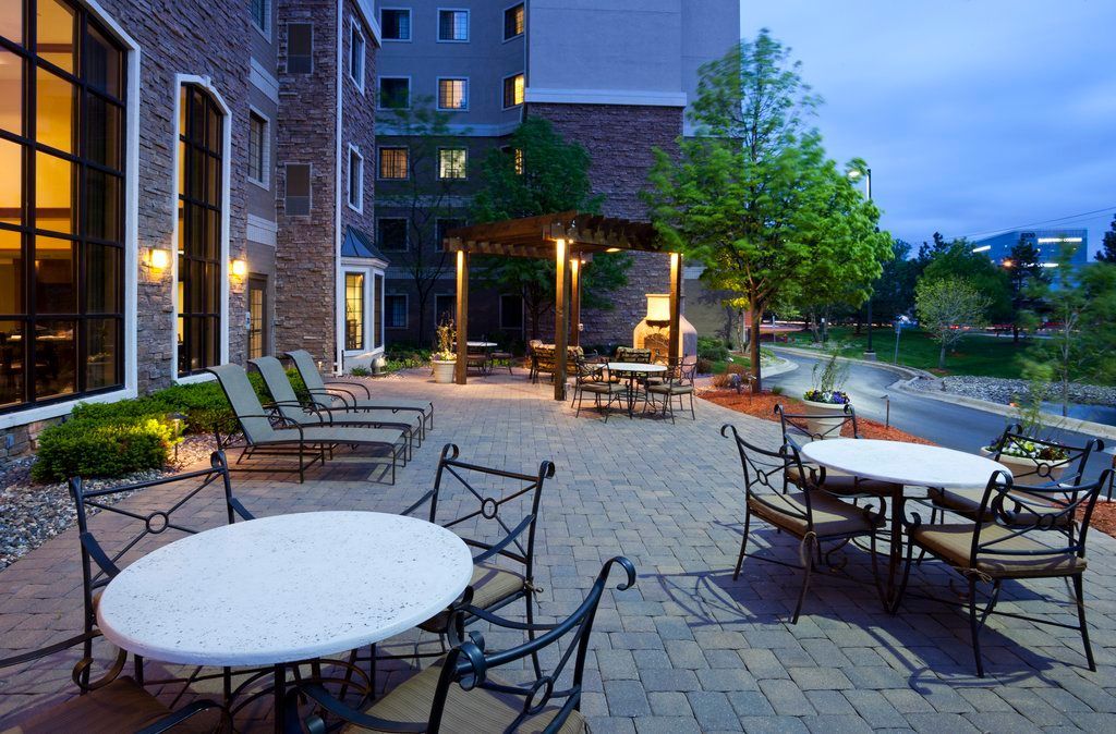 An outdoor patio with tables and chairs, a pergola, and a brick building with large windows.