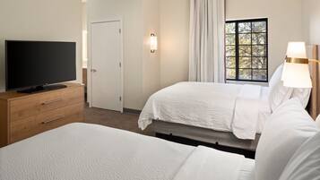 Ein Hotelzimmer mit Fernseher auf einer Holzkommode, zwei Betten mit weißer Bettwäsche, einem Fenster mit Vorhängen und einer Wandlampe.
