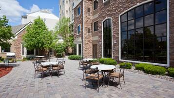 Une terrasse extérieure avec un sol en briques, entourée d’un bâtiment en pierre doté de grandes fenêtres et d’une porte.