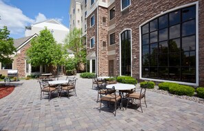 An outdoor patio with brick flooring, surrounded by a stone building with large windows and a door.
