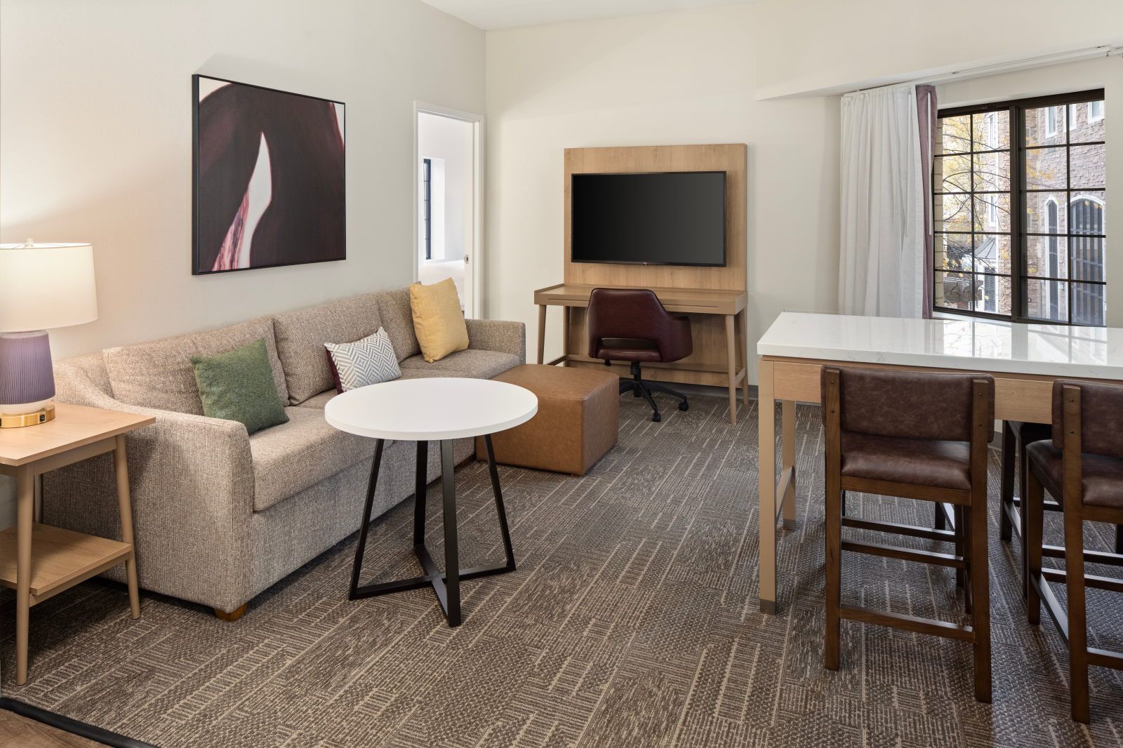 Une chambre d’hôtel avec un canapé, une table ronde, un téléviseur fixé sur un panneau en bois et une fenêtre avec des rideaux.