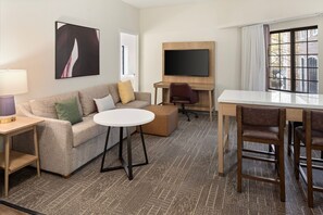A hotel room with a sofa, a round table, a TV mounted on a wooden panel, and a window with curtains.