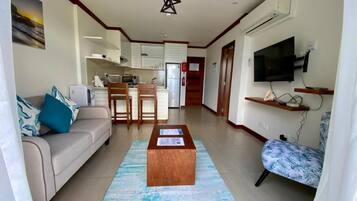 Una sala de estar con un sofá, una mesa de centro y un televisor. Se observa una cocina al fondo con armarios y un microondas.