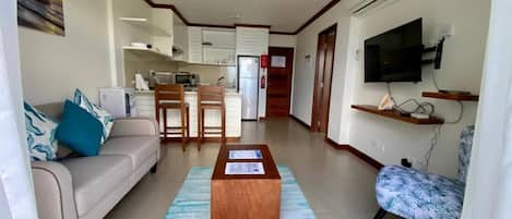 Una sala de estar con un sofá, una mesa de centro y un televisor. Se observa una cocina al fondo con armarios y un microondas.